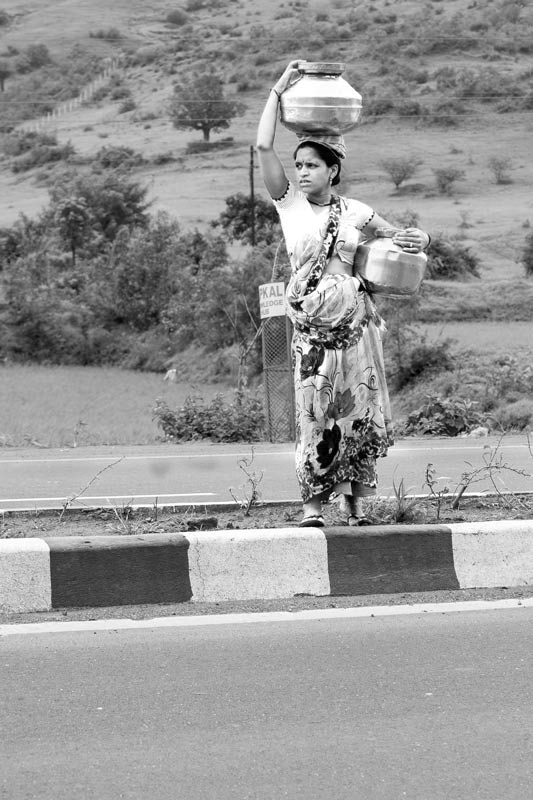 Rural Drinking Water Challenges - A pregnant rural woman carrying multiple loads of drinking water from a well across a busy motorable highway in Nashik district, Maharashtra